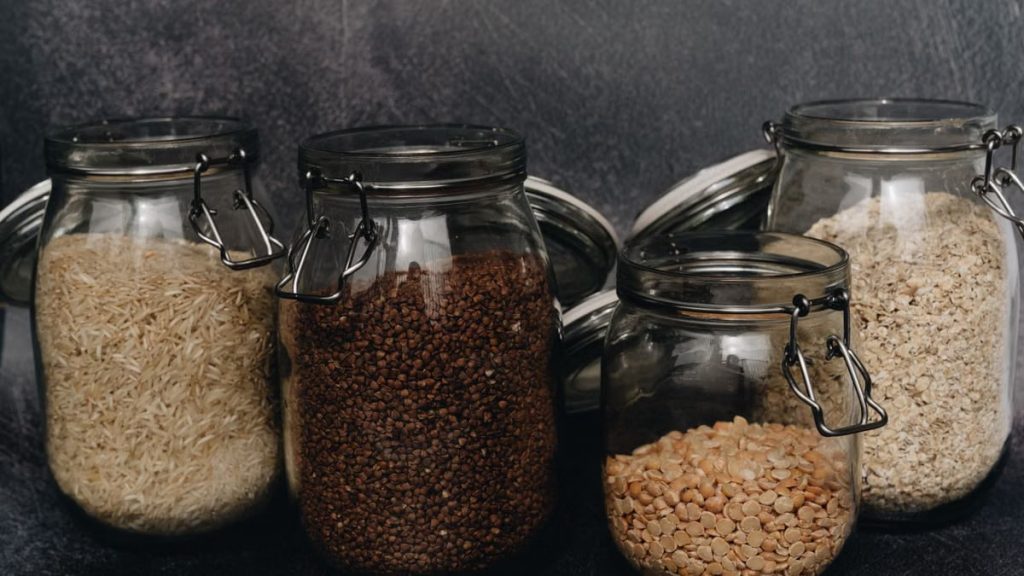 Four glass jars filled with buckwheat, lentils, rice, and oats on a dark surface, ideal for pantry organization.